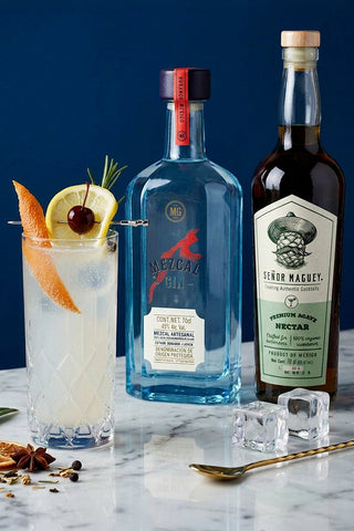Two bottles of Melfitz Gin and Sevier Macquet Nectar with a cocktail garnished with citrus and a cherry on a marble surface.