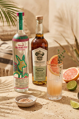 Two bottles of tequila and a cocktail with grapefruit and lime on a sandy surface.